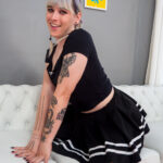 Tranny with thick tattooed arms poses on bed wearing a skirt.